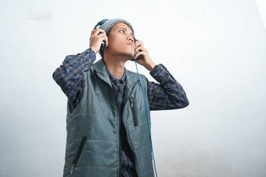 asian young man wearing dark blue vest and beanie wearing headphones, isolated on white background focused on listening to music via smartphone.