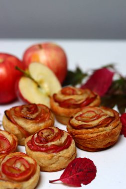 Gül şeklinde elmalı kekler, kırmızı elmalar, beyaz arka planda sonbahar yaprakları. Tarçınlı ve ballı elmalı turtaların en üstteki fotoğrafı.. 