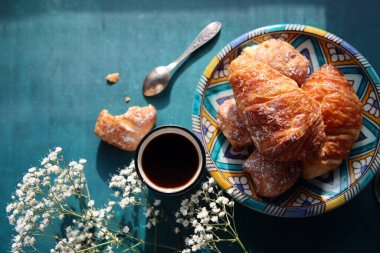 Taze pişmiş kruvasan, koyu mavi arka planda sade kahve ve çigila çiçekleri. Güzel bir sabah fotoğrafı. Yemek natürmort. 