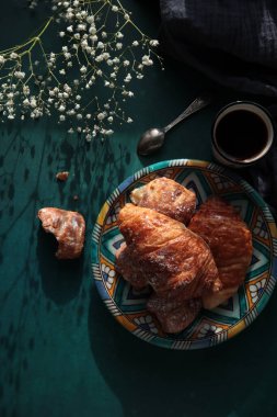 Taze pişmiş kruvasan, koyu mavi arka planda sade kahve ve çigila çiçekleri. Güzel bir sabah fotoğrafı. Yemek natürmort. 