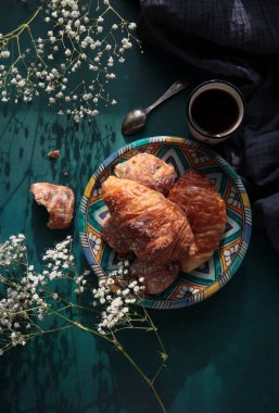 Taze pişmiş kruvasan, koyu mavi arka planda sade kahve ve çigila çiçekleri. Güzel bir sabah fotoğrafı. Yemek natürmort. 