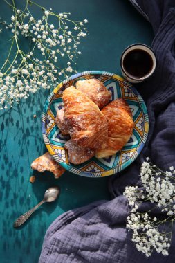 Taze pişmiş kruvasan, koyu mavi arka planda sade kahve ve çigila çiçekleri. Güzel bir sabah fotoğrafı. Yemek natürmort. 