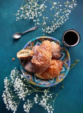 Taze pişmiş kruvasan, koyu mavi arka planda sade kahve ve çigila çiçekleri. Güzel bir sabah fotoğrafı. Yemek natürmort. 