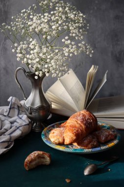 Yeni pişmiş kruvasanlarla dolu bir hayat, açık bir kitap, klasik gümüş vazoda beyaz çigila çiçekleri. Hafta sonu kahvaltısı konsepti. 