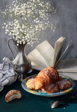 Yeni pişmiş kruvasanlarla dolu bir hayat, açık bir kitap, klasik gümüş vazoda beyaz çigila çiçekleri. Hafta sonu kahvaltısı konsepti. 