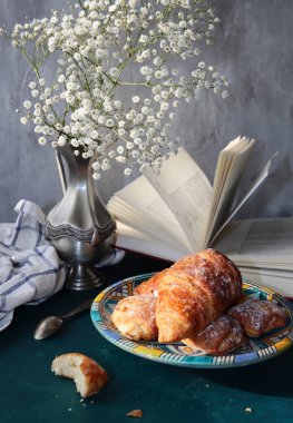 Yeni pişmiş kruvasanlarla dolu bir hayat, açık bir kitap, klasik gümüş vazoda beyaz çigila çiçekleri. Hafta sonu kahvaltısı konsepti. 