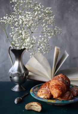 Yeni pişmiş kruvasanlarla dolu bir hayat, açık bir kitap, klasik gümüş vazoda beyaz çigila çiçekleri. Hafta sonu kahvaltısı konsepti. 