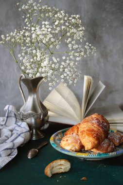 Yeni pişmiş kruvasanlarla dolu bir hayat, açık bir kitap, klasik gümüş vazoda beyaz çigila çiçekleri. Hafta sonu kahvaltısı konsepti. 