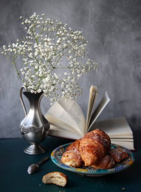 Yeni pişmiş kruvasanlarla dolu bir hayat, açık bir kitap, klasik gümüş vazoda beyaz çigila çiçekleri. Hafta sonu kahvaltısı konsepti. 
