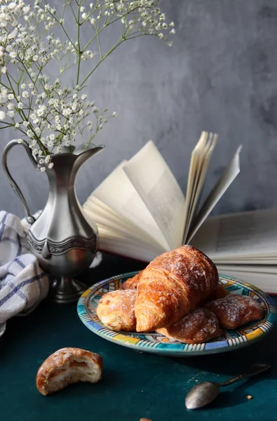 Yeni pişmiş kruvasanlarla dolu bir hayat, açık bir kitap, klasik gümüş vazoda beyaz çigila çiçekleri. Hafta sonu kahvaltısı konsepti. 