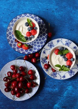 Yoğurt kâseleri ve tatlı kiraz kasesiyle dolu renkli bir hayat. Kopya alanı olan mavi desenli arkaplan. Vejetaryen kahvaltısı yakın plan fotoğrafı. Yaz meyveleri yakın.. 