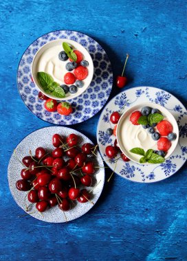 Yoğurt kâseleri ve tatlı kiraz kasesiyle dolu renkli bir hayat. Kopya alanı olan mavi desenli arkaplan. Vejetaryen kahvaltısı yakın plan fotoğrafı. Yaz meyveleri yakın.. 