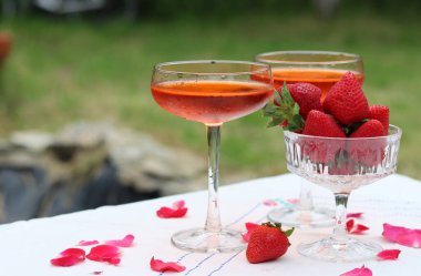 Bardakta kokteyl ve bahçe masasında taze çilek. Arkaplanda yeşil ağaç yaprakları var. Yaz akşamı. 