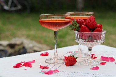 Bardakta kokteyl ve bahçe masasında taze çilek. Arkaplanda yeşil ağaç yaprakları var. Yaz akşamı. 