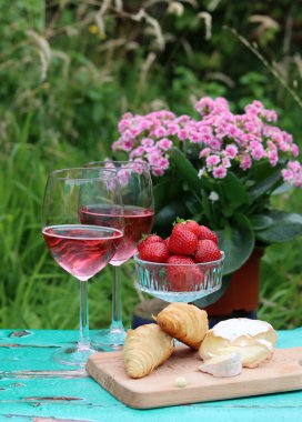 İki bardak gül şarabı, pembe çiçekler, çilek, peynir ve taze kruvasan. Bahçede piknik. Masada yaz yemeği. 