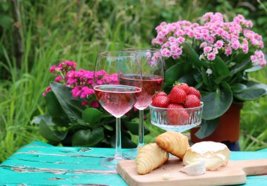 İki bardak gül şarabı, pembe çiçekler, çilek, peynir ve taze kruvasan. Bahçede piknik. Masada yaz yemeği. 