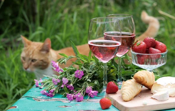 Summer romantic picnic. Sparkling wine, fruit and flowers on a wooden table. Ginger cat on a background. Selective focus.