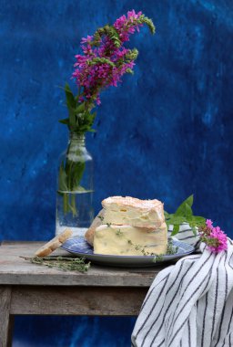 Rustic still life with flowers in a bottle, cheese with honey and herbs. Blue textured background with copy space. Food still life photo. 