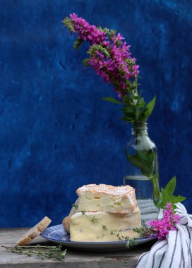 Rustic still life with flowers in a bottle, cheese with honey and herbs. Blue textured background with copy space. Food still life photo. 