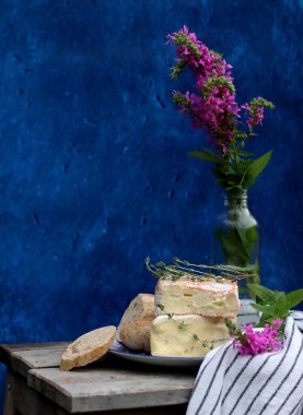 Rustic still life with flowers in a bottle, cheese with honey and herbs. Blue textured background with copy space. Food still life photo. 