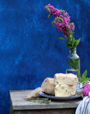 Rustic still life with flowers in a bottle, cheese with honey and herbs. Blue textured background with copy space. Food still life photo. 