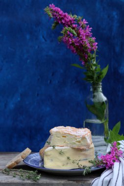 Rustic still life with flowers in a bottle, cheese with honey and herbs. Blue textured background with copy space. Food still life photo. 