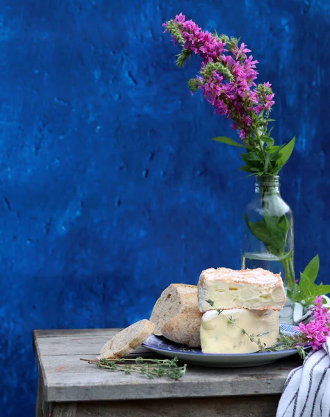 Rustic still life with flowers in a bottle, cheese with honey and herbs. Blue textured background with copy space. Food still life photo. 