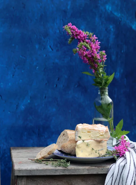 Rustic still life with flowers in a bottle, cheese with honey and herbs. Blue textured background with copy space. Food still life photo.