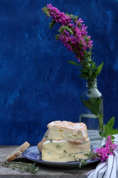 Rustic still life with flowers in a bottle, cheese with honey and herbs. Blue textured background with copy space. Food still life photo. 