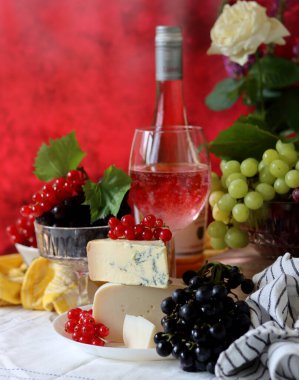 Wine, cheese, fruit and flowers on a table. Vibrant colors of fresh berries and grapes. Colorful still life with glass of pink wine, red and white grapes, gorgonzola cheese and red currant. 