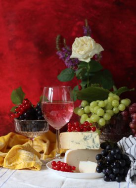 Wine, cheese, fruit and flowers on a table. Vibrant colors of fresh berries and grapes. Colorful still life with glass of pink wine, red and white grapes, gorgonzola cheese and red currant. 