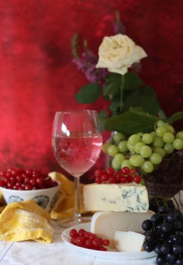 Wine, cheese, fruit and flowers on a table. Vibrant colors of fresh berries and grapes. Colorful still life with glass of pink wine, red and white grapes, gorgonzola cheese and red currant. 