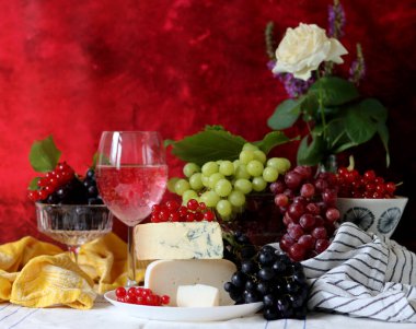 Wine, cheese, fruit and flowers on a table. Vibrant colors of fresh berries and grapes. Colorful still life with glass of pink wine, red and white grapes, gorgonzola cheese and red currant. 