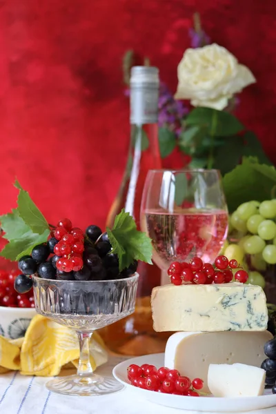 Wine, cheese, fruit and flowers on a table. Vibrant colors of fresh berries and grapes. Colorful still life with glass of pink wine, red and white grapes, gorgonzola cheese and red currant. 