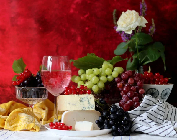 Wine, cheese, fruit and flowers on a table. Vibrant colors of fresh berries and grapes. Colorful still life with glass of pink wine, red and white grapes, gorgonzola cheese and red currant. 