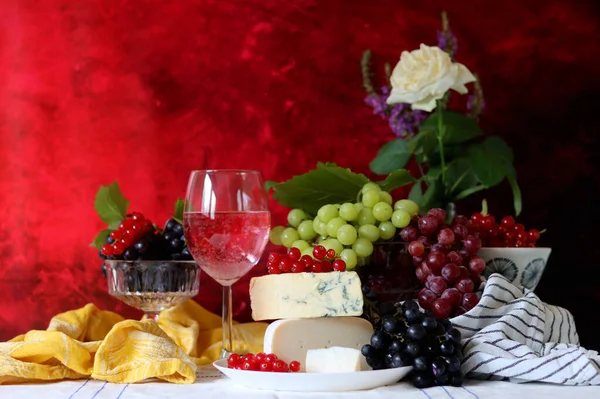Wine, cheese, fruit and flowers on a table. Vibrant colors of fresh berries and grapes. Colorful still life with glass of pink wine, red and white grapes, gorgonzola cheese and red currant. 