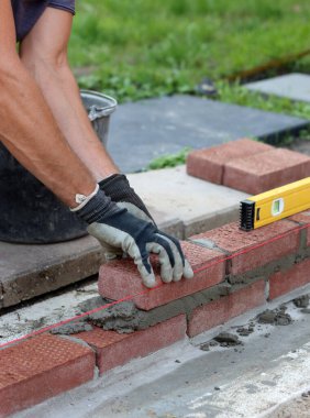 Male hands in protective gloves holding red bricks. Construction worker builds masonry.  Brickwork close up photo. 