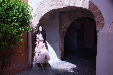 Orta yaşlı, kıvırcık saçlı, somon rengi bir takım giyen, güneş ışınlarını alırken duvar kemerine yaslanan İspanyol bir kadın. Moda, güzellik, güneş, şehir kavramı.