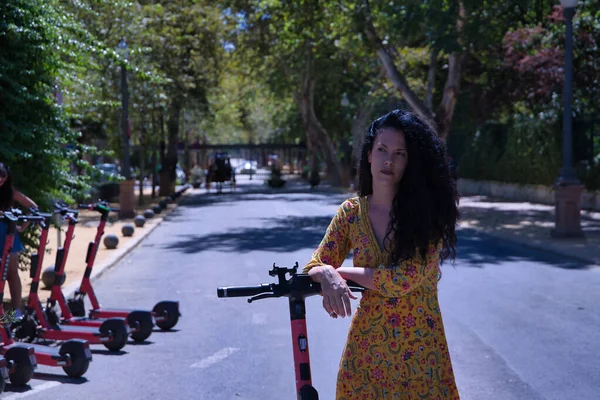 Middle-aged adult Hispanic woman with black curly hair, wearing a colorful outfit, leaning on an electric scooter. Concept scooter, urban mobility, electricity, battery, ecology.