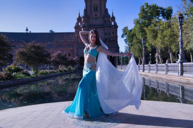 Orta yaşlı, turkuaz elbiseli, yapay elmaslı İspanyol kadın, beyaz peçeli göbek dansı. Göbek dansı konsepti.