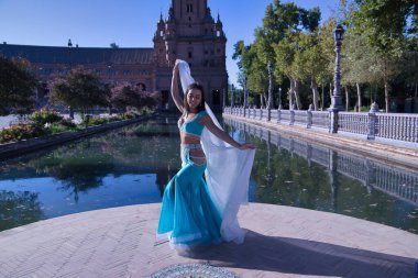 Orta yaşlı, turkuaz elbiseli, yapay elmaslı İspanyol kadın, beyaz peçeli göbek dansı. Göbek dansı konsepti.