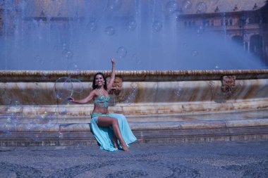 Orta yaşlı İspanyol kadın, su yeşili bir elbise giyiyor, yapay elmasları var, göbek dansı için, etrafında sabun köpükleriyle çeşmede oturuyor. Göbek dansı konsepti.