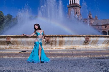 Orta yaşlı İspanyol kadın, turkuaz kostümlü ve yapay elmaslı, göbek dansı için, bir gökkuşağının oluştuğu bir çeşmenin önünde dans ediyor. Göbek dansı konsepti.