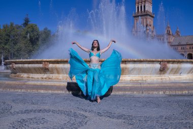 Orta yaşlı İspanyol kadın, turkuaz renkli kostümlü ve yapay elmaslı, göbek dansı yapmak için, elbisesini bir gökkuşağının oluştuğu bir çeşmenin önünde kaldırıyor. Göbek dansı konsepti.