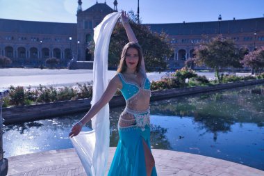 Orta yaşlı, turkuaz elbiseli, yapay elmaslı İspanyol kadın, beyaz peçeli göbek dansı. Göbek dansı konsepti.