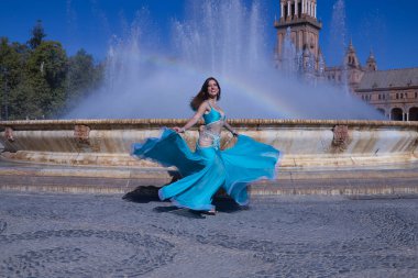 Orta yaşlı İspanyol kadın, turkuaz kostümlü ve yapay elmaslı, göbek dansı için, bir gökkuşağının oluştuğu bir çeşmenin önünde dans ediyor. Göbek dansı konsepti.