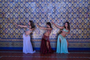 Orta yaşlı, güzel renkli ve boncuklu elbiselerle göbek dansına giden üç İspanyol kadın, arka planda güzel fayanslardan oluşan bir duvarla dans ederken figürler yapıyorlar. Göbek dansı konsepti.
