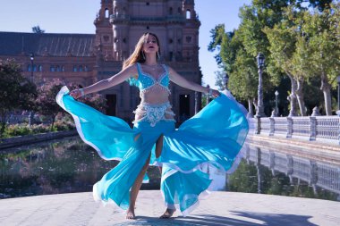 Orta yaşlı, turkuaz elbiseli, yapay elmasları olan, göbek dansı yaparken elbisesini tutan İspanyol bir kadın. Göbek dansı konsepti.