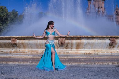 Orta yaşlı İspanyol kadın, turkuaz kostümlü ve yapay elmaslı, göbek dansı için, bir gökkuşağının oluştuğu bir çeşmenin önünde dans ediyor. Göbek dansı konsepti.