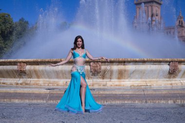 Orta yaşlı İspanyol kadın, turkuaz kostümlü ve yapay elmaslı, göbek dansı için, bir gökkuşağının oluştuğu bir çeşmenin önünde dans ediyor. Göbek dansı konsepti.